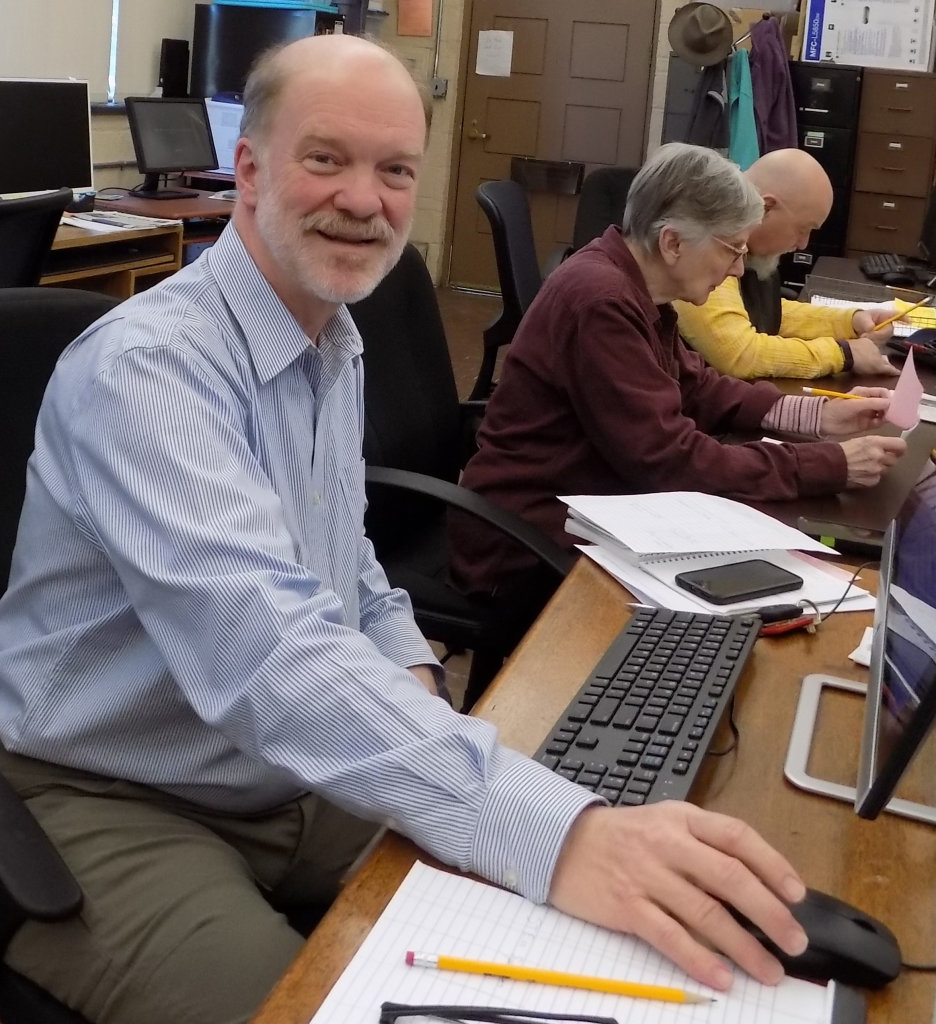 Gary Childs smiles while seated at a computer in the News Review office, reaching out to use a mouse. Behind him, two other people sit at a table, reading.