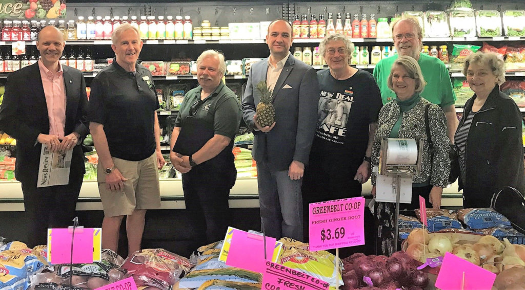 Eight people stand in front of the cold case in the co-op's produce department. In front of them is a display of bagged potatoes and onions with pink price signs.