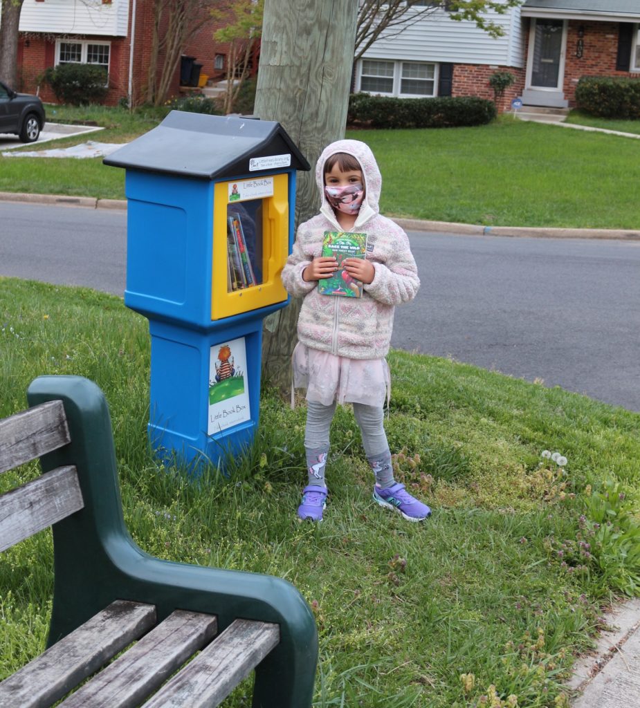 A girl holds a book to her chest. She is standing next to a blue plastic box on a blue pillar. A black roof covers the box. It has a yellow door with a window, through which more books are visible.