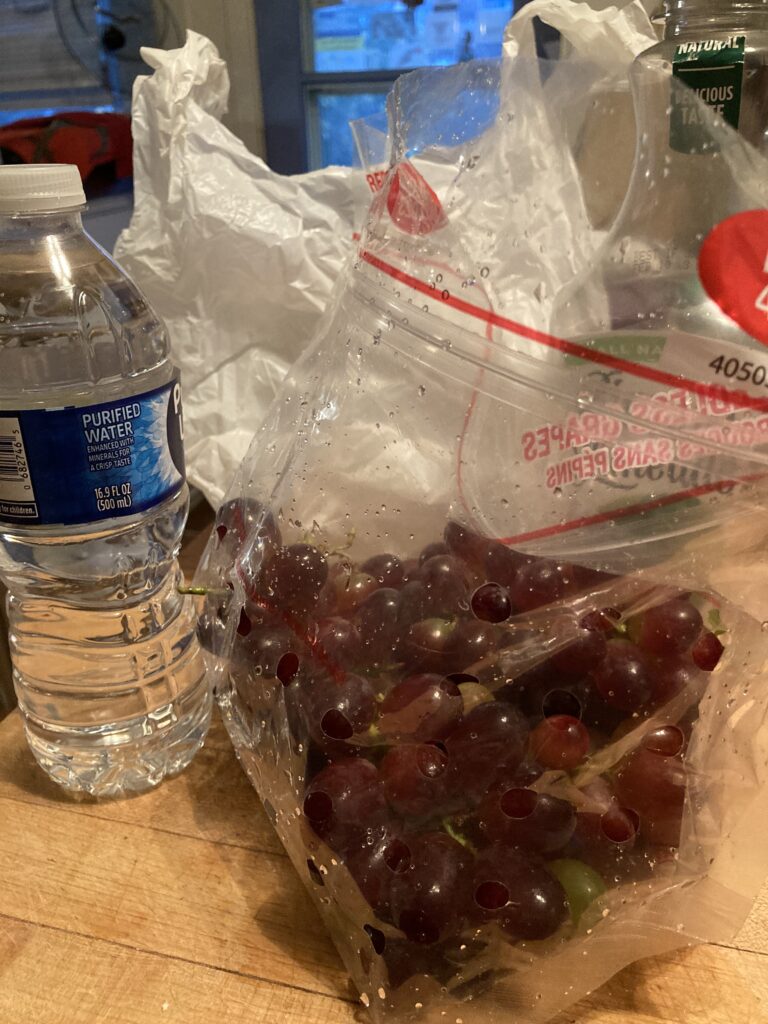 Purple grapes in a clear plastic bag, a single-use plastic water bottle with a blue label, and an opaque white plastic grocery bag sit on a table.