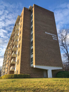 A brown 8-story building stands against a blue sky with light cloud cover, next to a green lawn.