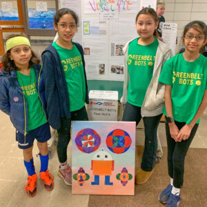 Four children, all wearing green T-shirts that say Greenbelt Bots in yellow letters. They stand around a colorful project. 