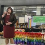A dark-haired person in a crimson knee-length dress and black jacket holds a microphone and sings, in front of a table with rainbow streamers and a green sign reading "Emerald City Pride In Memory of Ric Gordon."