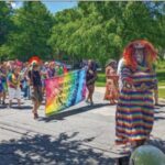 A person wearing a rainbow-striped dress and curly orange wig smiles with an outstretched hand, leading the parade down a suburban street among trees and yards. Behind them, two people carry a tie-dyed sign reading Greenbelt Honk! Situation. Behind them are parade participants in brightly colored clothing.