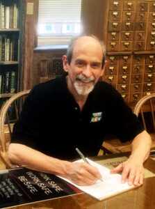 Chris Cherry, wearing a black polo shirt with the Greenbelt Recreation logo, sits at a table, writing on a notepad, and smiles for the camera. Behind him are bookshelves and a card catalog cabinet. 