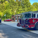 A red fire engine is parked outside the long brown garage with white doors. Two people stand in the parking lot in the background.