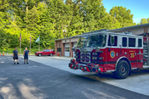 A red fire engine is parked outside the long brown garage with white doors. Two people stand in the parking lot in the background.