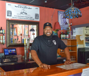 A black man wearing a black polo shirt and baseball cap stands behind the bar, with his hands on the polished wood bar top. To his right, a blue glass hanging lamp. Behind him, the bar taps and a computer terminal for ordering.