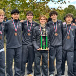 Seven young men stand side by side, wearing dark-colored shirts and slacks, with medals on multicolored ribbons around their necks. The center student holds a trophy with three green pillars.