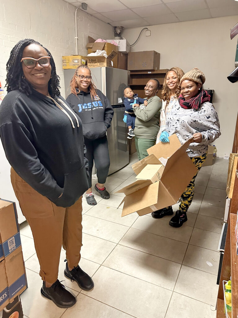 A dark-haired, dark-skinned person wearing a black sweatshirt, brown slacks, and light-colored glasses smiles for the camera in the foreground of an off-white room with off-white tiled floor. Four other people (plus one baby held in arms) stand behind them. One of those people holds a large cardboard box.