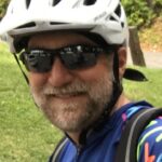 A light-skinned man wears a white bike helmet, sunglasses, and a blue shirt with bright designs. He has a close-trimmed pale beard and mustache. He is outside, with grass and a tree in the background.