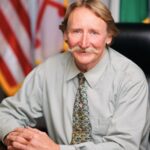 Rodney Roberts, a blond light-skinned man with a handlebar mustache, wears a light gray-green shirt with a dark green patterned tie. He sits at a table in front of the U.S. flag, the Maryland flag, and the Greenbelt flag.