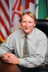 Rodney Roberts, a blond light-skinned man with a handlebar mustache, wears a light gray-green shirt with a dark green patterned tie. He sits at a table in front of the U.S. flag, the Maryland flag, and the Greenbelt flag.