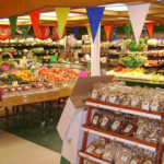 The co-op grocery store's produce department. Colorful produce and packages line shelves on two walls of the space, while more colorful produce rests on an island in the middle. At right, a display holds bags of packaged items. Colorful triangle banners hang from the ceiling.