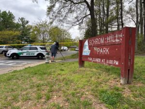 A brown sign on a grassy field bears the words SCHROM HILLS PARK in white, plus the Greenbelt city logo. Next to it, police cars park in a parking lot, behind a banner of yellow tape. 