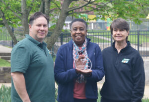 Three people stand side by side: a white man with brown hair, in green, a Black woman with short dark hair, in blue and pink, and a white woman with short brown hair, in black. The Black woman holds a round clear glass award with engraved writing.