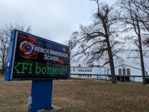 A DKFI school sign with the school name and cardinal logo. A digital component reads KFI bonjour! 