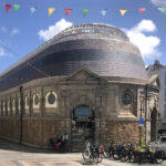 A long narrow building rounded at one end. Cobblestones pave the bottom half, with fancy brickwork alternating with ornate windows on the top half. A sloping grey tiled roof with a band of decorative blue tiles. Skylights at the top. The "coop" logo on the front door. Bike parking out front.