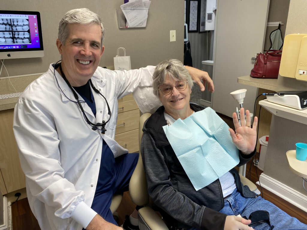 A seated dentist with one arm across the back of the patient chair. The patient, wearing the paper dropcloth, smiles and waves.