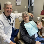 A seated dentist with one arm across the back of the patient chair. The patient, wearing the paper dropcloth, smiles and waves.