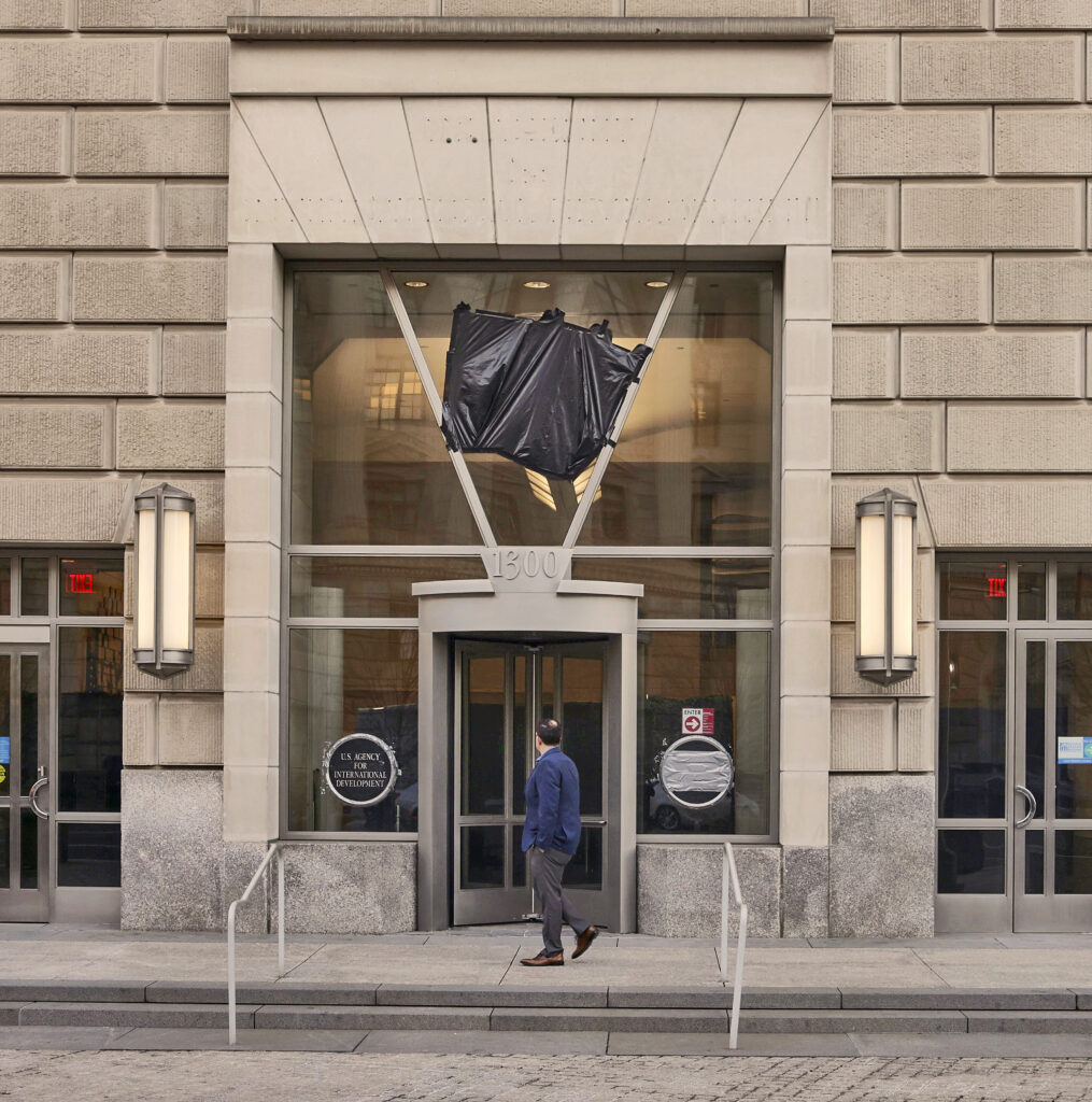 A beige stone building with a large glass window surrounding the door. Black tape covers a sign above the door. In the stonework above the glass, rows of rivet holes show where lettering was removed. A person walks past along the sidewalk.