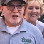 A light-skinned couple stand together and smile. Marc, in front, wears a baseball cap, glasses, and a gray polo shirt with the Greenbelt Recreation logo in green. Diane, behind him, has light hair.