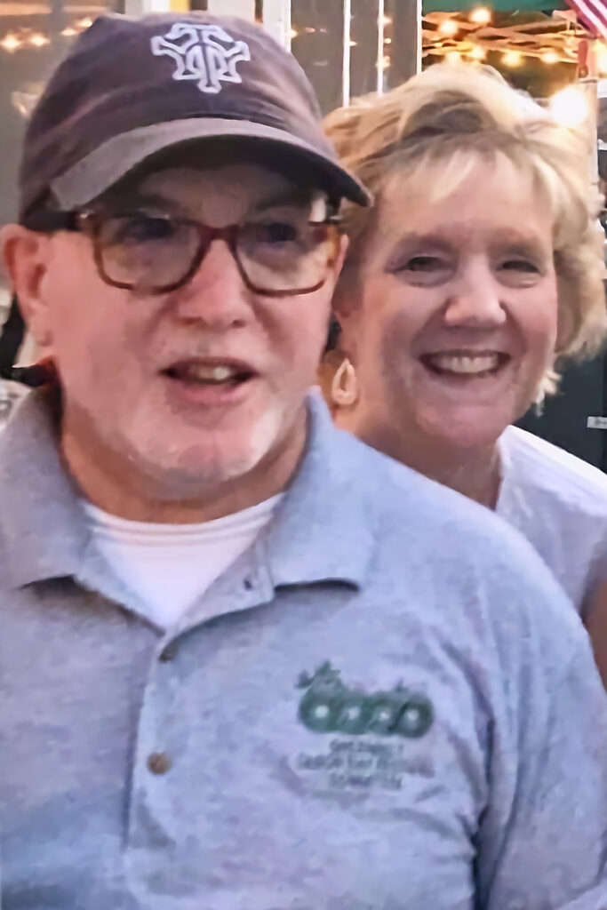 A light-skinned couple stand together and smile. Marc, in front, wears a baseball cap, glasses, and a gray polo shirt with the Greenbelt Recreation logo in green. Diane, behind him, has light hair.