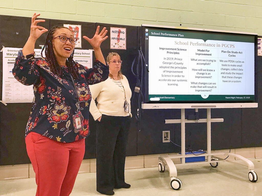 Foreground: a light-brown-skinned woman, with long dark hair, glasses, a black floral shirt and red pants. She raises both hands, fingers splayed. Her mouth is open and eyes wide in an expression of eagerness. Behind her, a blond, light peach skinned woman in a white blouse and black pants stands next to a projector screen, with a neutral posture and expression. The image on screen begins, "School Performance Plan: School Performance in PGCPS".