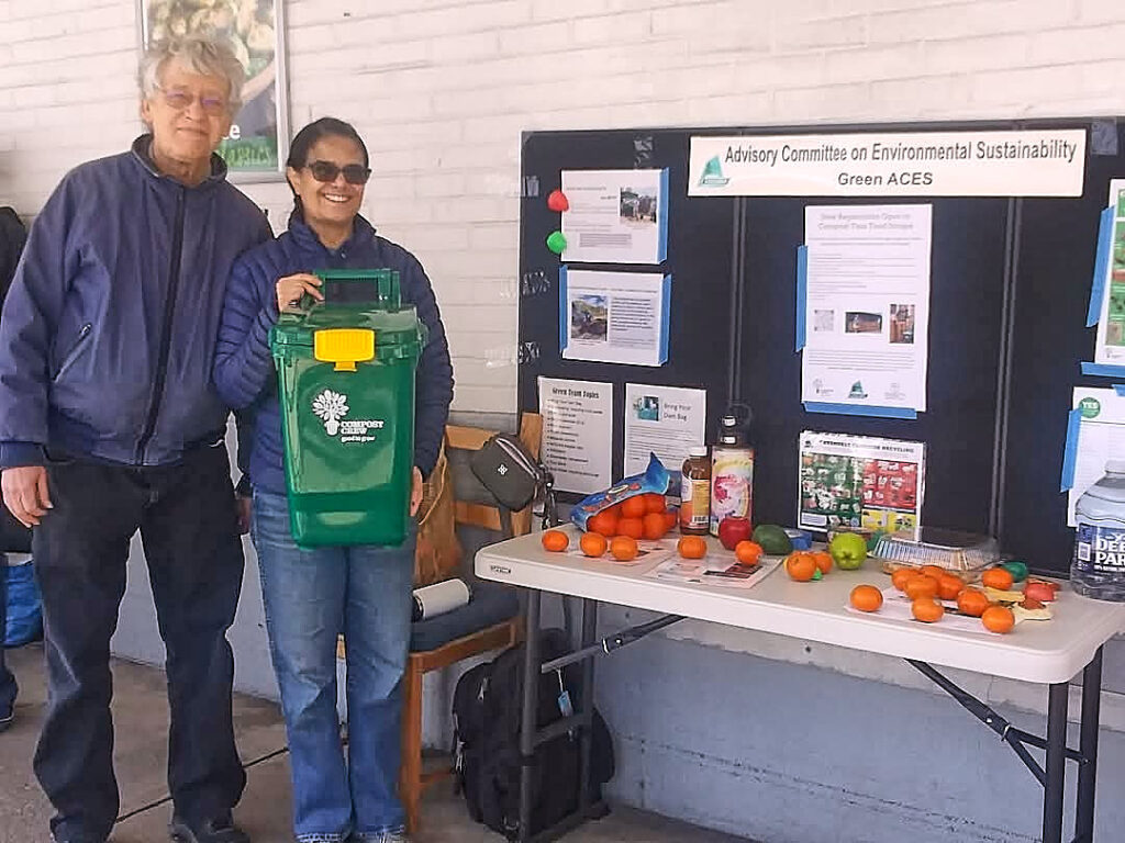 Two people stand together at left. One holds a small green plastic bin with a yellow clasp. They stand next to a table with a black display board that has white informational panels on it. On the table in front of the board are oranges, other fruits, and plastic containers.